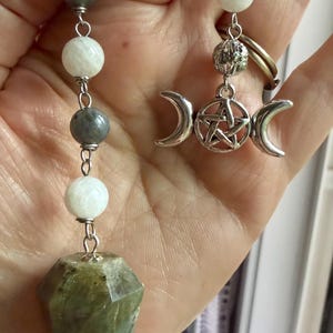 May include: A pendulum featuring a faceted green stone point suspended from a silver-toned chain. The chain includes alternating white and gray spherical beads, a small textured silver bead, and a silver-toned triple moon pentacle charm.