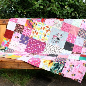 Könnte beinhalten: Eine Patchwork-Decke aus verschiedenen bunten Stoffen in Rosa, Rot, Grün und Gelb. Die Decke besteht aus Quadraten und liegt auf einer Holzbank.