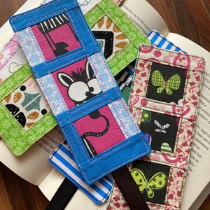 Könnte beinhalten: Eine Sammlung handgefertigter Stoff-Lesezeichen mit farbenfrohen Designs. Jedes Lesezeichen zeigt ein einzigartiges Muster, darunter ein Klavier, ein Kaninchen und ein Schmetterling. Die Lesezeichen sind aus verschiedenen Stoffen und Farben gefertigt, einige mit Bändern.