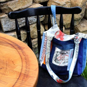 Könnte beinhalten: Eine blaue und weiße Patchwork-Tragetasche mit einer Karte von Marrakesch auf der einen Seite und dem Text "This is the way" auf der anderen Seite. Die Tasche liegt auf einem schwarzen Stuhl vor einer Steinmauer.