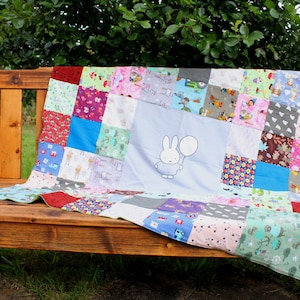 Könnte beinhalten: Ein bunter Patchwork-Quilt mit einem zentralen Feld, das ein Cartoon-Kaninchen mit einem Ballon zeigt. Der Quilt besteht aus verschiedenen gemusterten Stoffen in Blau-, Rosa-, Grün- und Rottönen. Er ist über eine Holzbank drapiert.