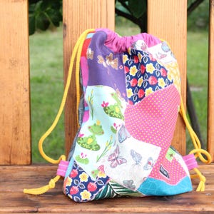 Könnte beinhalten: Eine bunte Kordeltasche aus Patchworkstoff mit verschiedenen Mustern, darunter Frösche, Schmetterlinge, Blumen und Punkte. Die Tasche hat gelbe Kordeln und einen rosa Verschluss.