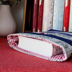 Könnte beinhalten: Ein Buchcover mit roter und weißer karierter Innenausstattung. Das Cover ist aus blau-weiß gestreiftem Stoff mit weißem Hintergrund und einem Muster aus schwarzen und weißen Illustrationen. Das Buchcover ist über ein weißes Buch gefaltet.