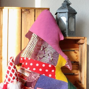 Könnte beinhalten: Ein Patchwork-Weihnachtsbaumkissen mit rosa Spitze und verschiedenen gemusterten Stoffen in Rot, Lila, Gelb und Grün. Daneben sitzt ein kleiner Wichtel mit roter und weißer Mütze und Bart. Im Hintergrund eine dekorative Laterne.
