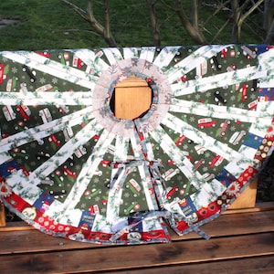 Könnte beinhalten: Ein Weihnachtsbaumrock mit festlichem Design. Der Rock hat einen grünen Hintergrund mit weißen Streifen und roten, weißen und blauen Akzenten. Er hat eine kreisförmige Öffnung in der Mitte und ist mit einem roten Rand versehen. Der Rock hat einen Durchmesser von etwa 122 cm.