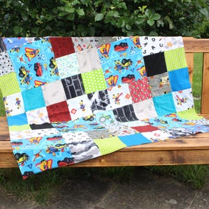 Könnte beinhalten: Eine bunte Patchwork-Decke in Blau-, Grün- und Rottönen. Die Decke besteht aus verschiedenen Stoffen mit unterschiedlichen Mustern, darunter ein Ziegelmuster, ein grün-weißes Karomuster und ein blaues Muster mit einem Baufahrzeug-Design. Die Decke ist auf einer Holzbank ausgestellt.