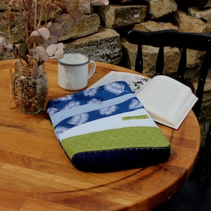 Könnte beinhalten: Ein gesteppter Buchumschlag mit blauem Palmenblattmuster, hellblauen und weißen Streifen und einem grünen, gemusterten Boden. Auf einem Holztisch mit einer Tasse, einem Buch und einem Glas getrockneter Blumen.