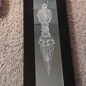 May include: A black framed mirror with a white etched design of a dagger with a crown and multiple faces on the handle.