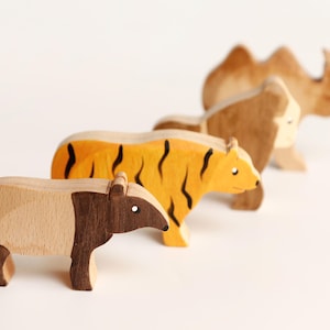 Puede incluir: Un conjunto de cuatro figuras de animales de madera dispuestas en fila. Las figuras incluyen un tapir de madera clara y oscura, un tigre amarillo con rayas negras, un gorila marr&oacute;n y un camello marr&oacute;n claro con dos jorobas en la espalda.