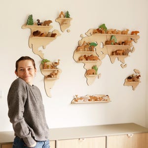 Może przedstawiać: Drewniane półki ścienne w kształcie mapy świata z różnymi drewnianymi figurkami zwierząt. Półki są zamontowane na białej ścianie. Półki mają kształt kontynentów i wysp. Zwierzęta obejmują niedźwiedzie, słonie i żyrafy.
