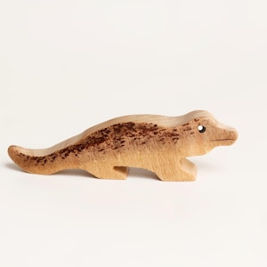Puede incluir: Una peque&ntilde;a figura de madera tallada a mano que representa un cocodrilo o caim&aacute;n. La pieza presenta un acabado de madera natural en tono beige claro con manchas o marcas moteadas de color marr&oacute;n oscuro en el lomo y la cola, y un ojo perforado.