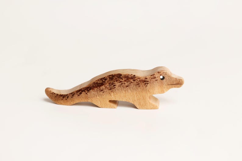 Puede incluir: Una peque&ntilde;a figura de madera tallada a mano que representa un cocodrilo o caim&aacute;n. La pieza presenta un acabado de madera natural en tono beige claro con manchas o marcas moteadas de color marr&oacute;n oscuro en el lomo y la cola, y un ojo perforado.
