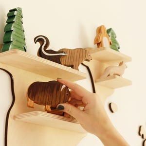 May include: Wooden wall shelf in a map design, with multiple shelves displaying wooden animal figurines. The animals include a skunk, bear, fox, and a musk ox. A green painted wooden tree is also on the shelf.