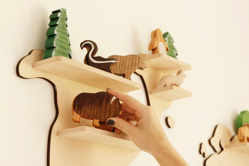 May include: Wooden wall shelf in a map design, with multiple shelves displaying wooden animal figurines. The animals include a skunk, bear, fox, and a musk ox. A green painted wooden tree is also on the shelf.