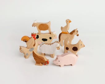 Puede incluir: Una colección de figuras de animales de granja de madera que incluye una vaca, un cerdo, una oveja, un caballo, una cabra, un gallo, una gallina, un gato y un perro. Las figuras presentan acabados de madera natural con detalles pintados.