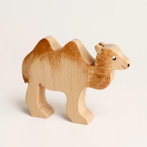 Puede incluir: Una figura de camello de madera con un acabado de veta de madera clara natural y detalles pintados en marr&oacute;n en las jorobas, el cuello y la cabeza. La figura tiene una silueta tallada con un peque&ntilde;o punto negro para el ojo y una l&iacute;nea.