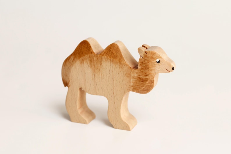 Puede incluir: Una figura de camello de madera con un acabado de veta de madera clara natural y detalles pintados en marr&oacute;n en las jorobas, el cuello y la cabeza. La figura tiene una silueta tallada con un peque&ntilde;o punto negro para el ojo y una l&iacute;nea.