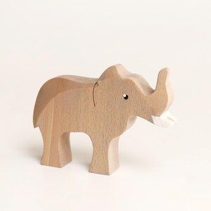 Puede incluir: Una figura de elefante de madera de color marr&oacute;n claro con un acabado liso, que presenta un peque&ntilde;o ojo pintado de negro y un colmillo pintado de blanco. La figura animal se muestra en posici&oacute;n de pie con una trompa curva y cuerpo redondeado.