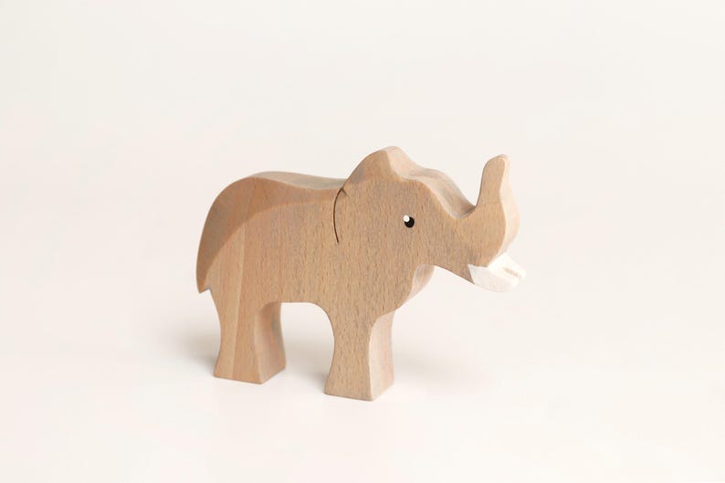 Puede incluir: Una figura de elefante de madera de color marr&oacute;n claro con un acabado liso, que presenta un peque&ntilde;o ojo pintado de negro y un colmillo pintado de blanco. La figura animal se muestra en posici&oacute;n de pie con una trompa curva y cuerpo redondeado.