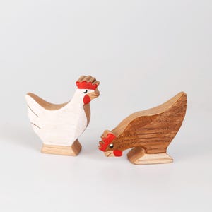 Op de afbeelding: Twee houten kippenbeeldjes. Eén is wit met bruine accenten, de andere is bruin. Beide hebben rode kammen en lellen. De kippen staan op een witte achtergrond.