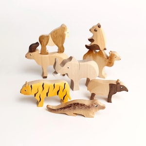 Puede incluir: Una colecci&oacute;n de siete figuras de animales de madera tallada que incluye un tigre amarillo con rayas negras, un rinoceronte de color claro, un camello, un oso, un gorila, un tapir y una criatura alargada, todos con acabados de madera natural.