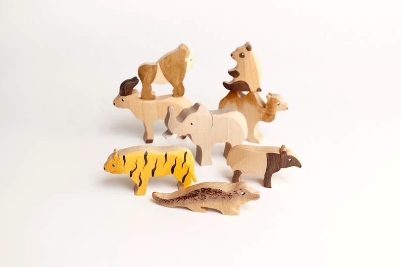 Puede incluir: Una colecci&oacute;n de siete figuras de animales de madera tallada que incluye un tigre amarillo con rayas negras, un rinoceronte de color claro, un camello, un oso, un gorila, un tapir y una criatura alargada, todos con acabados de madera natural.