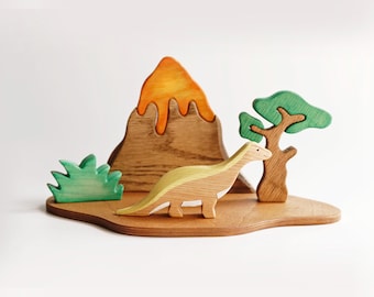 May include: Wooden dinosaur toy set featuring a volcano with orange lava, a green tree, and a dinosaur with yellow and green accents. The set is on a wooden base, creating a prehistoric scene.
