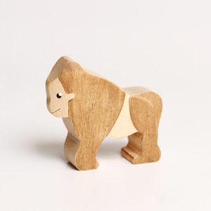 Puede incluir: Una figura de gorila de madera hecha de madera de tonos claros y medios. La figura animal presenta una superficie lisa y pulida con vetas de madera visibles, un ojo negro pintado simple y una l&iacute;nea curva que representa la boca.