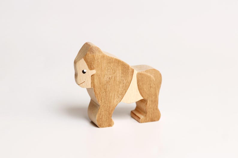 Puede incluir: Una figura de gorila de madera hecha de madera de tonos claros y medios. La figura animal presenta una superficie lisa y pulida con vetas de madera visibles, un ojo negro pintado simple y una l&iacute;nea curva que representa la boca.