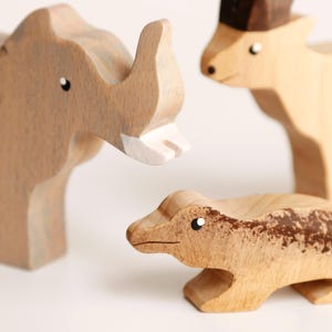 Puede incluir: Tres figuras de animales de madera que incluyen un elefante marr&oacute;n claro con un colmillo blanco, un ciervo beige con la cabeza marr&oacute;n oscuro y un lagarto marr&oacute;n claro con un patr&oacute;n texturizado en el lomo. Cada figura tiene ojos pintados.