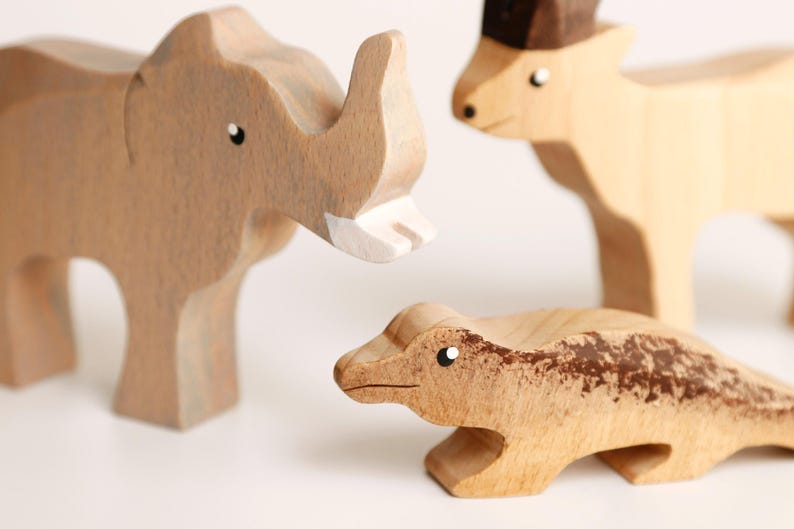 Puede incluir: Tres figuras de animales de madera que incluyen un elefante marr&oacute;n claro con un colmillo blanco, un ciervo beige con la cabeza marr&oacute;n oscuro y un lagarto marr&oacute;n claro con un patr&oacute;n texturizado en el lomo. Cada figura tiene ojos pintados.