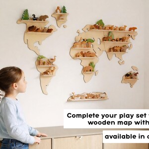 Puede incluir: Un mapa del mundo de madera de color claro para pared con estantes horizontales integrados. El mapa est&aacute; decorado con varias figuras peque&ntilde;as de madera de animales y &aacute;rboles. El texto en la imagen dice: Complete your play set with our wooden map with shelves available in our shop.