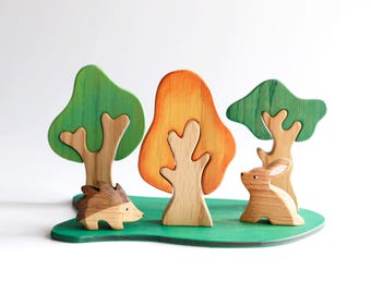 May include: Wooden toy set featuring a green, orange, and brown forest scene with a hedgehog and a rabbit. The set includes three trees and a base.