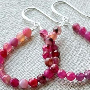 May include: A pair of hoop earrings featuring faceted, round beads in various shades of pink, magenta, and deep red. The beads are strung on a thin, silver-toned metal wire that forms a circular shape, finished with a simple silver-toned hook ear wire.