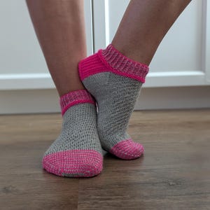 Könnte beinhalten: Ein Paar knöchelhohe Stricksocken mit einem hellgrauen, strukturierten Körper und leuchtend pinkfarbenen Bündchen sowie Zehenpartien. Die Socken haben ein geripptes Strickmuster am Knöchel und eine ausgeprägte Webstruktur im Fußbereich.