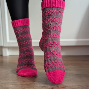 Peut inclure: Paire de chaussettes tricotées avec une base grise et un motif de losanges roses répété. Le bout et le bord sont roses unis. Les chaussettes sont présentées sur les jambes d'une personne, avec des leggings noirs visibles.