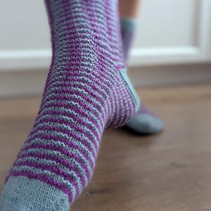 May include: A pair of hand-knitted socks featuring a horizontal striped pattern in alternating light blue and deep purple yarn. The socks have a solid light blue toe section, a matching light blue cuff, and a reinforced heel design in light blue.