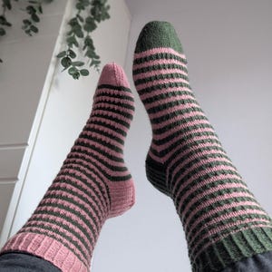 Könnte beinhalten: Nahaufnahme von zwei gestreiften Socken in Rosa und Grün. Die Socken haben einfarbige rosa Zehen und Fersen, mit grünen und rosa horizontalen Streifen auf dem Körper. Die Socken werden mit einer grauen Hose getragen.