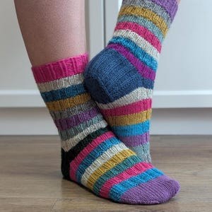 May include: A pair of knitted socks featuring a ribbed texture and horizontal stripes in vibrant colours including hot pink, mustard yellow, light blue, purple, grey, and black. The socks have a solid purple toe section and a dark blue heel section.