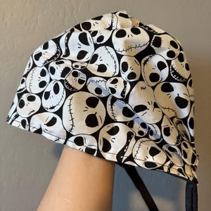 Puede incluir: Un sombrero con estampado en blanco y negro con un diseño repetido de la cara de Jack Skellington de Pesadilla antes de Navidad. El sombrero tiene una corbata negra y está hecho de una tela suave.