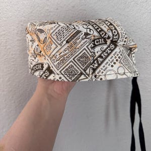 May include: A patterned surgical scrub cap with a black tie closure. The cap features a cream-colored background with brown and orange vintage-style text and illustrations. The text includes the words "Warts" and "Broomstick".