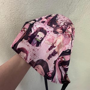 Peut inclure: Un bonnet rose à motifs avec des illustrations de personnages d'anime. Le bonnet présente un motif répété d'un personnage aux longs cheveux foncés et aux accents roses. Le bonnet a des liens noirs.