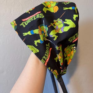 May include: Black fabric head covering featuring Teenage Mutant Ninja Turtles characters in green, orange, blue, and purple. The words "Teenage Mutant Ninja Turtles" are printed in white and red. The head covering has black ties.