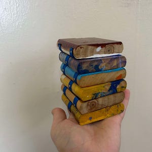 Könnte beinhalten: Ein Stapel kleiner, handbemalter Holzbücher in verschiedenen Farben, darunter Braun, Blau und Gelb. Jedes Buch hat ein blaues Band und wird in einer Hand gehalten. Die Bücher scheinen dekorativ zu sein oder zum Spielen.