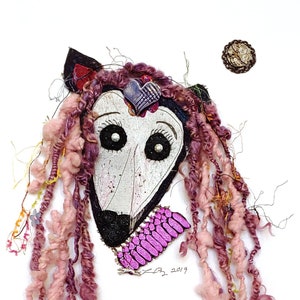 May include: A mixed media artwork featuring a whimsical animal face with pink yarn hair, black eyes, and a pink and white patterned collar. The artwork is signed and dated "L2 2019".