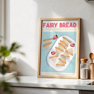 Puede incluir: Un póster enmarcado con la frase "FAIRY BREAD" en rosa. La imagen muestra sándwiches triangulares y fresas en una bandeja blanca sobre un fondo azul claro. El póster está en un marco de madera, expuesto sobre una encimera blanca.