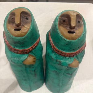 May include: Two ceramic figures with teal robes and tan faces. Each figure has a red beaded necklace and arms crossed at the chest. The figures have a distressed, aged appearance, suggesting a vintage or handcrafted origin.