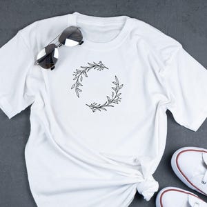Puede incluir: Camiseta blanca con un dise&ntilde;o simple de corona de flores negra.