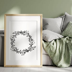 Puede incluir: Una impresi&oacute;n de corona floral en blanco y negro en un marco de madera claro, colgada en una pared sobre un sof&aacute; verde con almohadas.