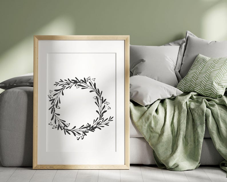 Puede incluir: Una impresi&oacute;n de corona floral en blanco y negro en un marco de madera claro, colgada en una pared sobre un sof&aacute; verde con almohadas.